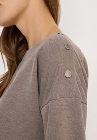 Brauner Strickpullover mit rundem Ausschnitt, strukturiertem Stoff und metallischen Akzenten an den Schultern sowie Knopfdetails.
