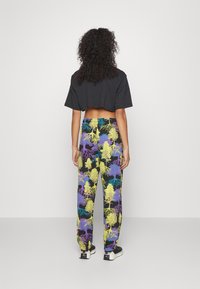Woman with curly hair wearing a black cropped t-shirt and colorful tree-patterned pants standing against a plain light background.
