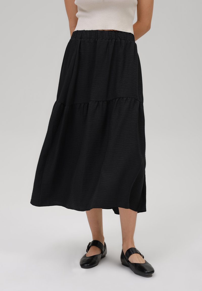 Jupe midi noire à volants avec une taille élastique, en tissu texturé et avec une coupe fluide, assortie à des chaussures Mary Jane noires.