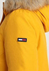 Gul vinterjacka med pälsfodrad huva. Har en svart, röd och vit logopatch på ärmen. Slät textur och modern design.