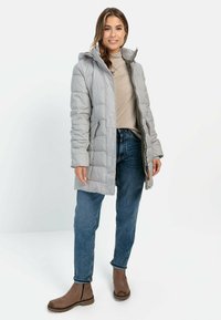 Polstret grå frakke med hætte, der har en frontlynlås, sidelommer og en afslappet pasform, parret med blå jeans og brune ankelstøvler.