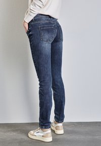 Jean en denim bleu foncé avec une coupe slim, doté d'une taille mi-haute, de deux poches arrière et d'une légère texture délavée. Associé à des baskets blanches et beiges.