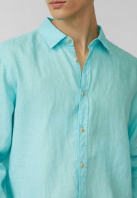 Camicia in lino azzurra chiara con bottoni in tonalità legno e colletto classico, che trasmette un aspetto morbido e testurizzato.