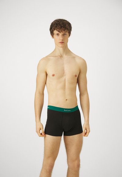 Caleçons pour hommes noirs avec une ceinture verte ; fabriqués en tissu lisse, présentant une étiquette de marque à l'avant. Modèle présenté dans une pose neutre.