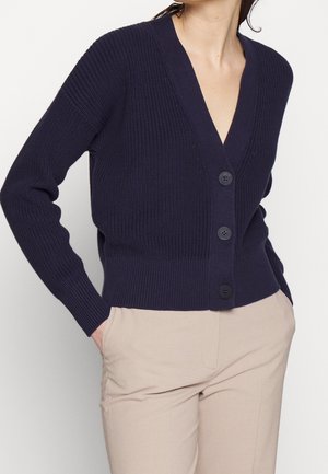 Žena oblečená v tmavě modrém zapínaném svetru s hlubokým výstřihem do V a béžových kalhotách, ruce v kapsách na jednobarevném pozadí.