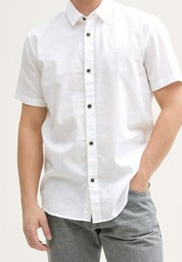 Weiße kurzärmlige Button-up-Hemd aus einem strukturierten Stoff, mit braunen Knöpfen und einem kleinen Logodetail auf der Brust. Kombiniert mit grauen Jeans.