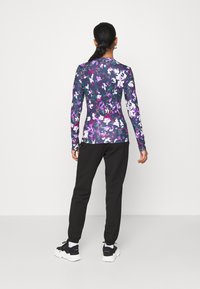 Långärmad åtsittande topp med ett abstrakt blommönster i lila, rosa och vitt; kombinerad med svarta joggingsbyxor och svarta träningsskor.