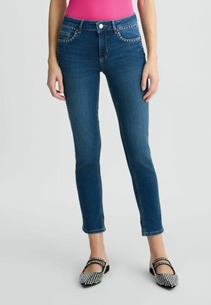 Jeans in denim con un design aderente, color blu scuro, borchie decorative sulle tasche e una lunghezza leggermente accorciata, abbinati a scarpe bianche e nere.