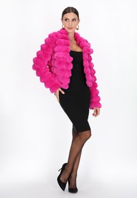 Vrouw in een felroze textuur bontjas over een fitted zwart jurkje, zwarte doorzichtige panty’s en zwarte hoge hakken, poseert met haar hand op haar heup.