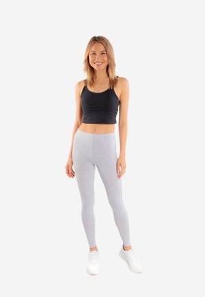 Kobieta uśmiechająca się, stojąca, ubrana w czarny krótki top, jasnoszare legginsy i białe trampki na jednolitym białym tle.