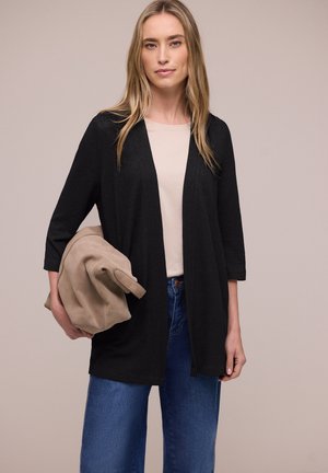 Donna che indossa un cardigan nero aperto, top beige, jeans blu, tiene una giacca beige piegata su uno sfondo beige uniforme.
