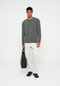 Mann in grauem Langarmshirt und weißen Hosen, der eine schwarze Tragetasche hält und vor einem weißen Hintergrund steht.