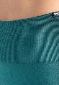 Turkooise geribbelde tailleband van een legging met gladde stof eronder. Voorzien van een klein zwart label met de tekst "SMILE" op de tailleband.