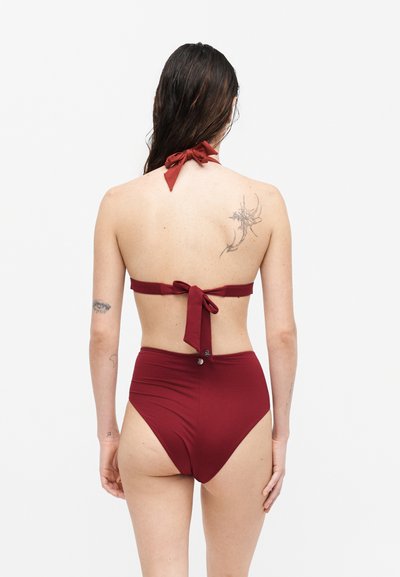 Γυναίκα με μακριά σκουρόχρωμα μαλλιά που φοράει ένα βουργουνδί μπικίνι δύο κομματιών με σλιπ ψηλόμεσο και δέσιμο στον λαιμό, δείχνοντας την πλάτη και τα τατουάζ.