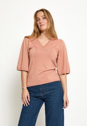 Ženska z dolgimi blond lasmi, oblečena v bleščečo prahasto roza majico z V-izrezom in napihnjenimi trikotnimi rokavi do komolcev ter modre kavbojke, stoji pred belim ozadjem.