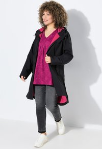 Manteau long noir avec des accents roses, doté d'un devant zippé, d'une capuche et de poches, porté sur un haut texturé violet et un jean gris.