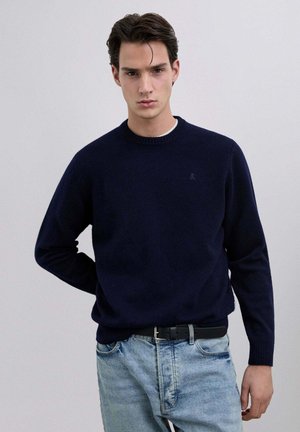 Suéter de punto azul marino con cuello redondo y puños acanalados. Combinado con jeans de mezclilla azul claro. Detalle sutil del logo en el pecho.