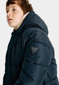 Marineblauwe pufferjack met capuchon, voorzien van een logo-patch op de linker schouder, gestikte details en een gladde, gestructureerde stofafwerking.