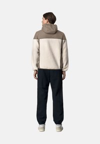 Giacca beige e tan foderata in pile con cappuccio, caratterizzata da un retro texturizzato e polsini elasticizzati, abbinata a pantaloni jogger neri e sneakers.