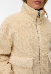 Beige, fluffig fleecejacka med hög krage, dragkedja framtill och två påsydda fickor. Tyget har ett strukturerat, mjukt utseende.