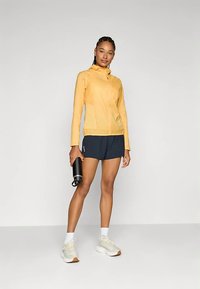 Viegls dzeltena krāsa jaka ar rāvējslēdzēja priekšpusi un kapuci, kopā ar tumši ziliem šortiem. Modelis tur melnu ūdens pudeli, nēsā ādas krāsas sporta apavus.