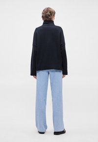 Suéter preto de gola alta com corte solto, mangas longas e bainha canelada, combinado com jeans de perna larga em azul claro e calçado preto.