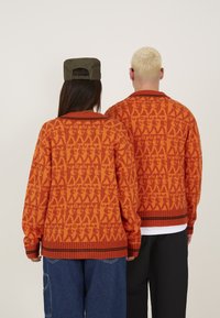 Pull en tricot orange présentant un motif répétitif, un col et des poignets côtelés. Porté par deux personnes avec un jean foncé et une tenue décontractée.