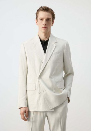 Junger Mann trägt cremefarbenen zweireihigen Anzug mit schwarzem Hemd mit Kragen darunter, steht vor einfachem hellem Hintergrund.
