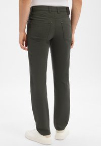 Man wearing dark olive straight-leg pants and white sneakers, standing with back facing the camera against a white background.