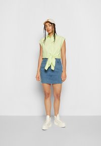 Camisa verde claro de mangas curtas atada na cintura, combinada com uma mini saia de ganga azul e tênis brancos de cano alto. Boné creme na cabeça.