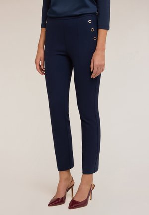 Pantalons navy ajustés en tissu lisse avec des détails latéraux présentant des œillets dorés, complétés par de élégantes talons bordeaux.
