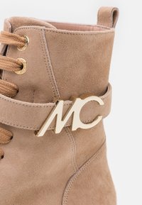 Bottine en suède beige à lacets avec logo en métal doré "MC", design à lacets et détail de surface texturé. Parfaite avec un lacet marron et un talon robuste.