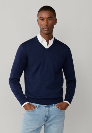 Mężczyzna w granatowym swetrze z dekoltem w serek, na białej koszuli z kołnierzykiem, w jasnoniebieskich dżinsach, stojący z rękami w kieszeniach na szarym tle.