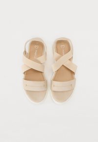 Sandales beige avec un design à lanières croisées, dessus en tissu doux, semelles rembourrées et semelle en caoutchouc blanc. Look minimaliste sans embellissements.