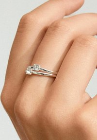 Anillo de plata con dos piedras preciosas transparentes, con un diseño suave y bandas delgadas que se entrelazan en una mano. Textura de piel suave visible.