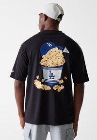 T-shirt en coton noir avec un motif de pop-corn dans un récipient rayé bleu et blanc, surmonté d'un couvercle bleu, imprimé au dos.