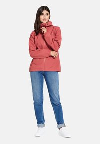 Waterdichte roze jas met een ritssluiting aan de voorkant, verstelbare capuchon en elastische manchetten, gecombineerd met blauwe jeans en witte sneakers.