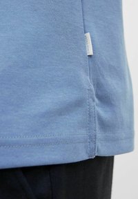 Šviesiai mėlynas medvilninis marškinėliai su tiesiu apdailos kraštu, šonine išpjova ir mažu prekės ženklo žymeku kairėje pusėje, arti siūlės.