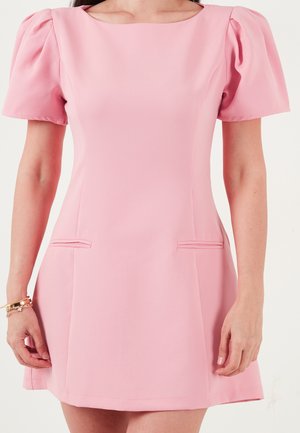 Mujer con un vestido corto rosa con mangas cortas abullonadas y dos pequeños detalles de bolsillos delanteros, de pie con los brazos relajados.