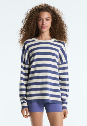 Jonge vrouw met krullend haar die een blauw-wit gestreepte lange mouwen trui en paarse korte broek draagt, staande tegen een effen achtergrond.