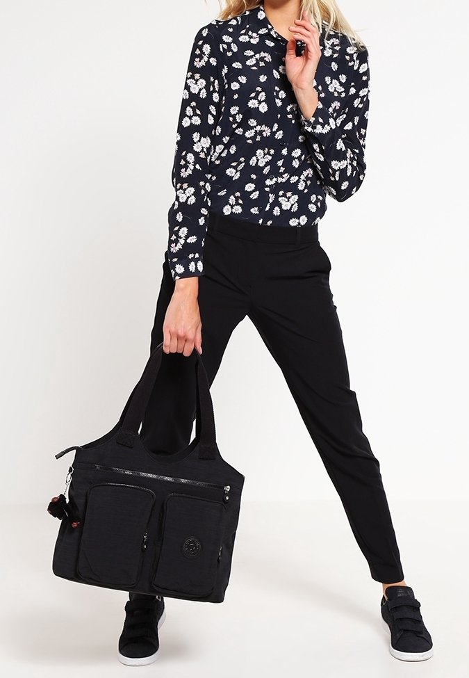 Femme portant un pantalon noir et une chemise noire à fleurs à manches longues, tenant un grand sac cabas noir avec plusieurs poches zippées.