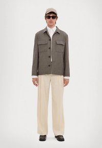 Braune karierte Jacke mit zwei Brusttaschen, kombiniert mit einem weißen Hemd und cremefarbenen Hosen mit weitem Bein. Accessoires umfassen eine beige Mütze und eine Sonnenbrille.