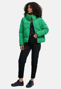 Giacca a vento verde con colletto alto, design trapuntato e chiusura frontale. Indossata sopra una maglietta nera e abbinata a pantaloni neri e sneakers.