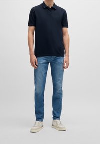 Marinblå polo med kort ärm och en liten broderad logotyp, i kombination med ljusblå jeans och krämfärgade sneakers.