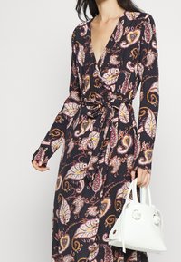 Žena v dlouhém černém šatě s paisley vzorem a vázačkou v pase, držící malou bílou kabelku s uzlovými detaily.