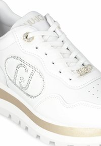 Sneaker in pelle bianca con un accento dorato sulla suola, caratterizzato da un logo in strass e un design con lacci e occhielli metallici.