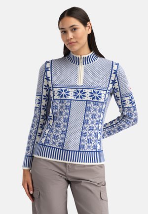 Kvinne iført en langermet blå og hvit mønstret genser med frontglidelås og beige cargobukser, poserer med en hånd bak ryggen.