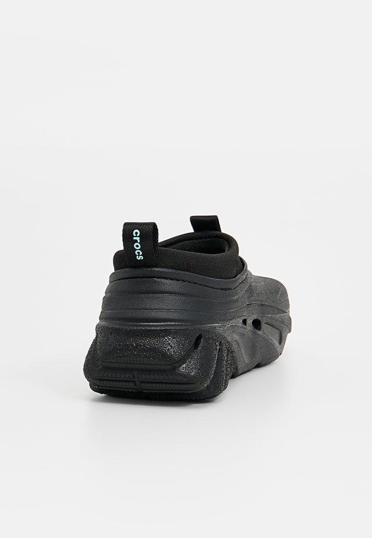 Zwarte instapper met een getextureerde bovenkant, rubberen zool en subtiele merklogo op de hiel. Heeft een verhoogde achterkant en gladde contouren.