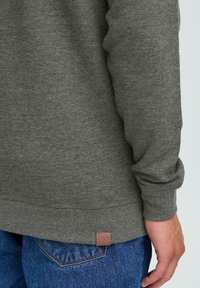 Grå hettegenser med strukturert stoff, lange ermer og en liten merket lærpatch på sømmen, kombinert med blå denimjeans.