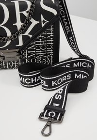 Borsa Michael Kors in bianco e nero con grande stampa del logo e tracolla nera staccabile con testo bianco Michael Kors e sistema di chiusura in metallo.